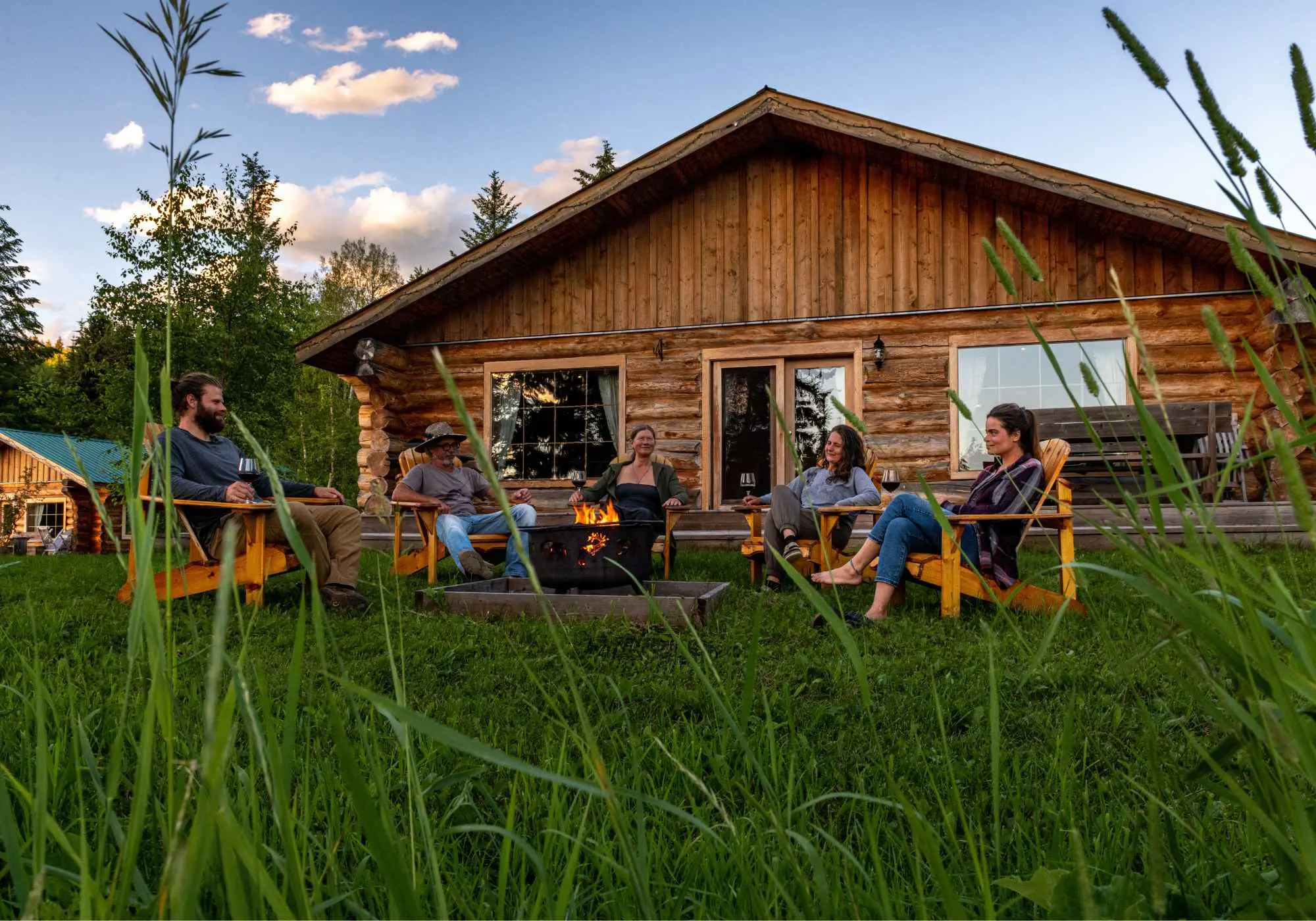 A small group sitting around a fire outside a log cabin at Kayanara in the evening, tall grasses in the foreground.