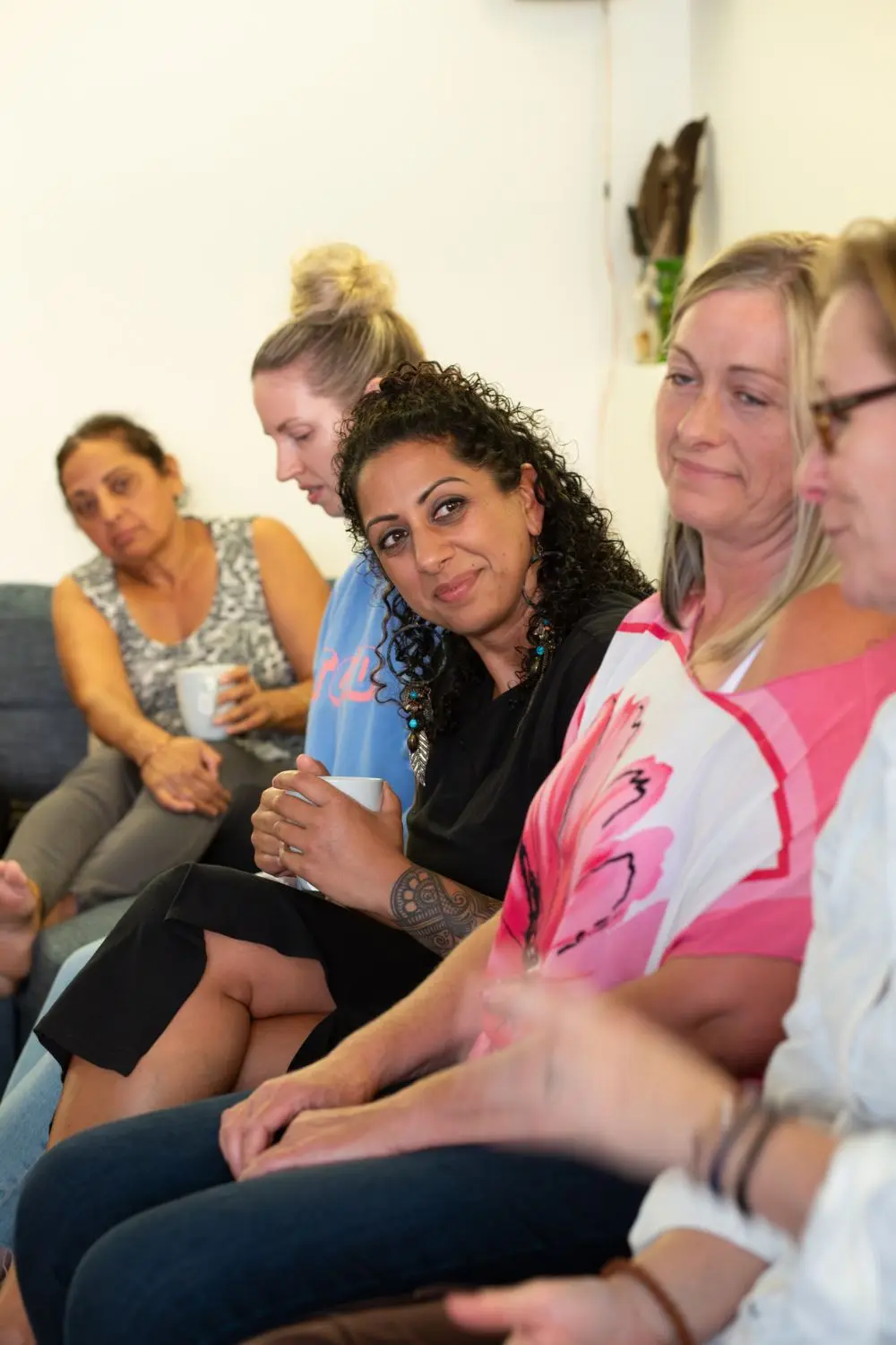 Pam Dhillon on a couch among a circle of women at a gathering, mugs in hand, listening.