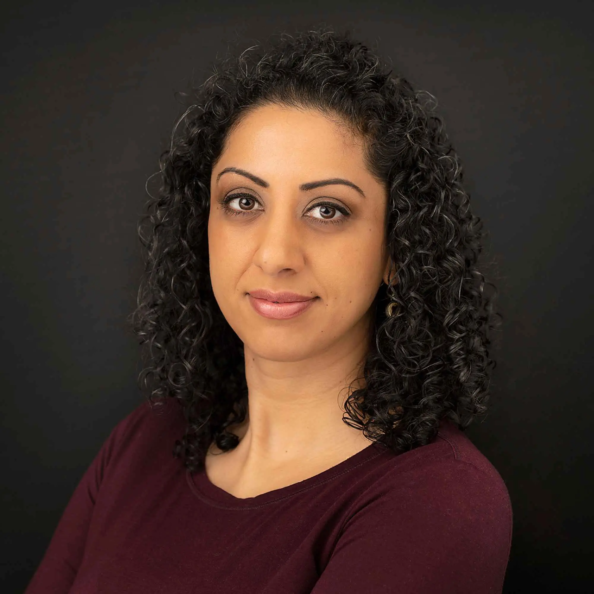 Pam Dhillon, looking directly at the camera, against a dark studio background.
