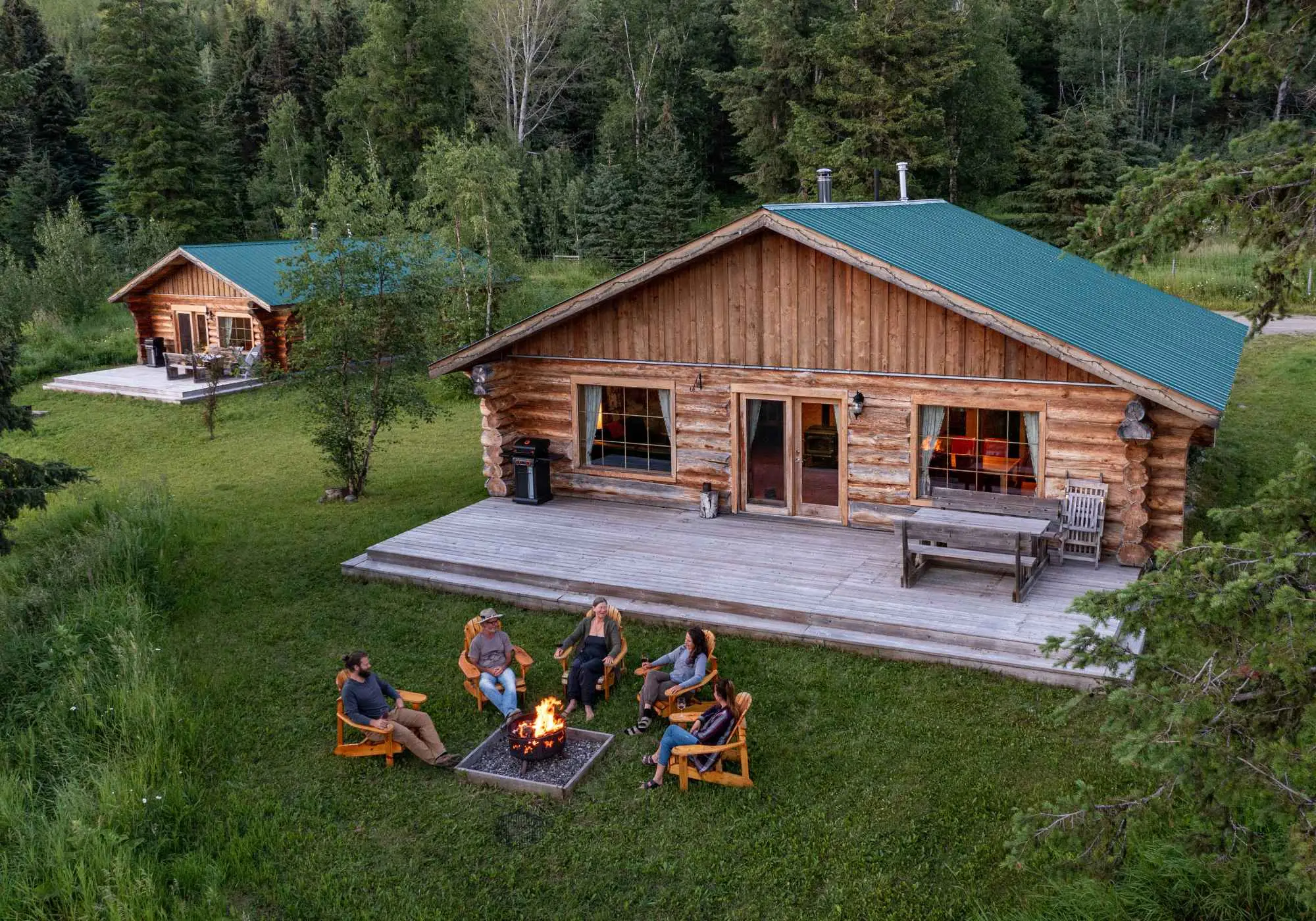 A handcrafted log cabin at Kayanara seen from above, surrounded by forest, with people gathered around a fire on the grass.